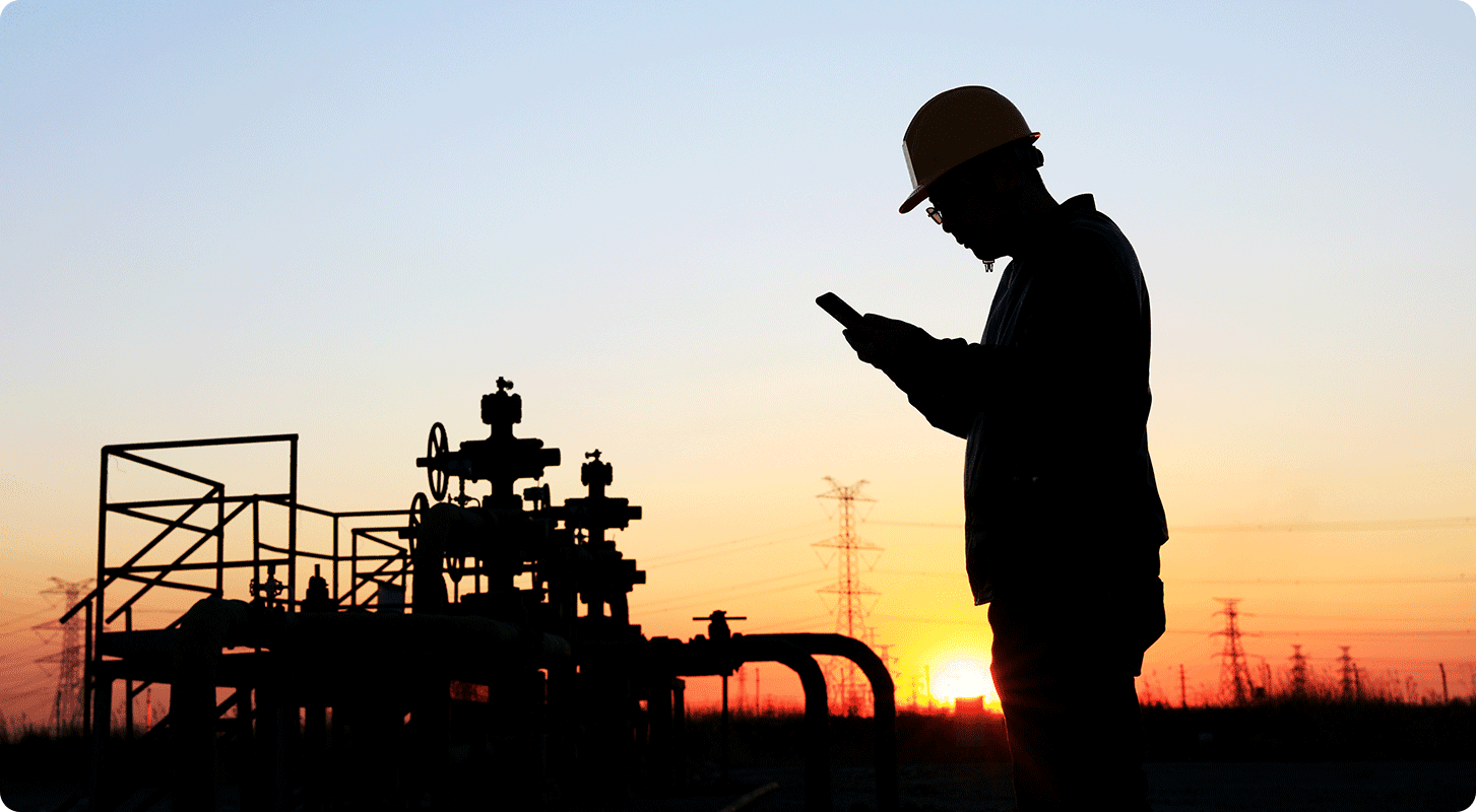 A silhouetted person in a hard hat checks a mobile device near industrial machinery at sunset.